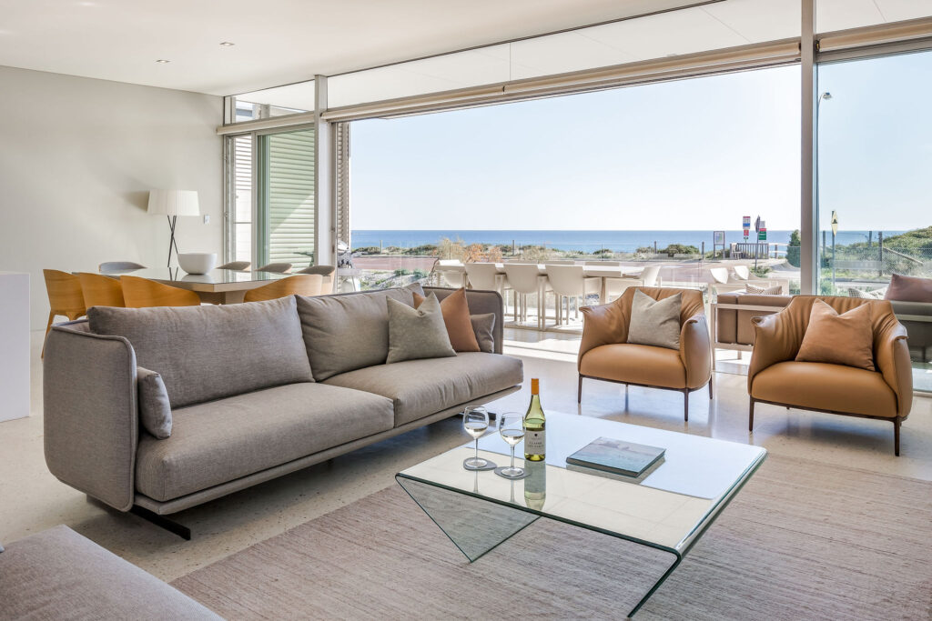 Grey couch and brown armchairs in foreground with balcony and ocean view in background.