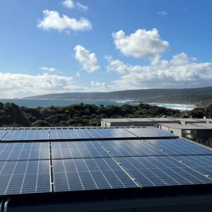 View of solar panels on a building with blue ocean and green foliage in the distance. 