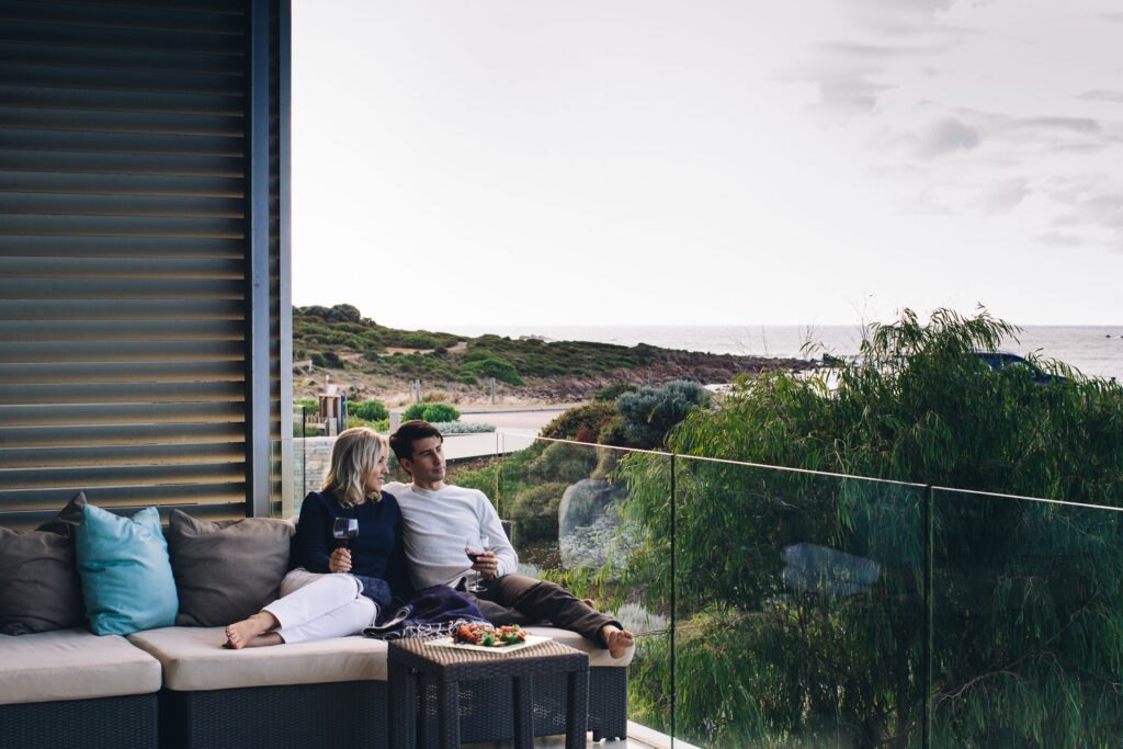 A couple relaxes on a balcony with wine and food. The ocean is visible in the background. 